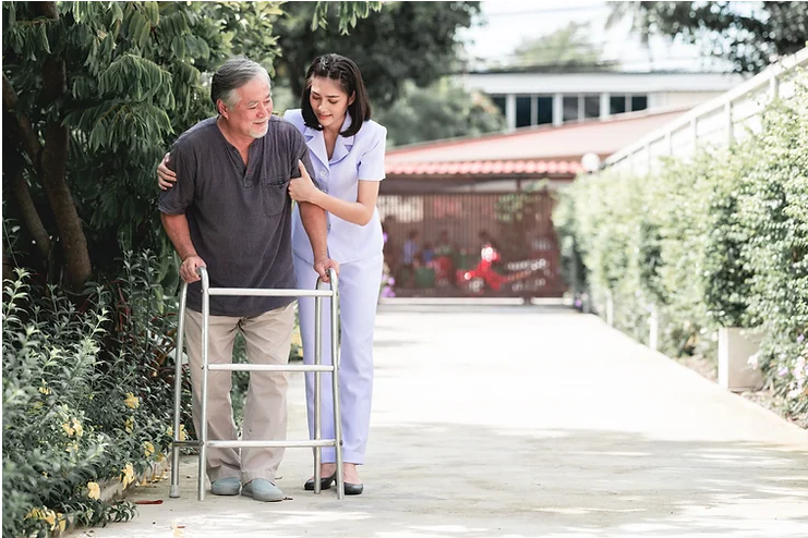 Parkinson’s-Disease-Causes-Symptoms-and-Treatments A nurse assists an elderly man with a walker, outdoors in a garden setting.